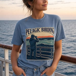 May include: A dusty blue short-sleeve t-shirt featuring a graphic print of a person looking through binoculars at the ocean from a deck. The text PELAGIC BIRDING is printed above the illustration, and FIRST LIGHT DECK WATCH is printed below.