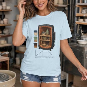 May include: A light blue t-shirt featuring a graphic of a pottery kiln with three shelves holding ceramic pieces, numbered color swatches, and a firing temperature graph with the text "RESULTS PENDING IN FIRE" printed below the kiln illustration.