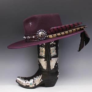 Peut inclure: Un chapeau prune à large bord, orné d'une bande décorative et d'une étoile. Il repose sur une botte de cowboy noire avec des éléments décoratifs argentés et un bout pointu. Des plumes et un tissu sombre ornent le chapeau.