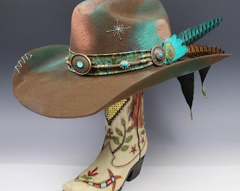 Puede incluir: Un sombrero de vaquero marrón con adornos turquesa y plateados, una estrella y una pluma. El sombrero se exhibe sobre una bota de vaquero color crema con diseños florales y geométricos.