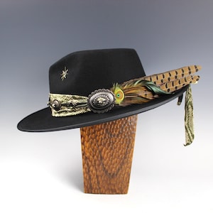 Può includere: Cappello fedora nero con fascia decorativa, accento a stella e abbellimenti di piume. Il cappello presenta una fascia color oro con un elemento centrale ovale argentato. Un gruppo di piume e un tessuto fantasia pendono dal lato.