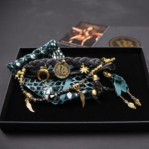 May include: A black box contains a collection of jewelry. A braided black bracelet with a gold coin and a crescent moon charm is visible. Other bracelets with gold beads and charms, and a teal fabric with a scale pattern are also present.
