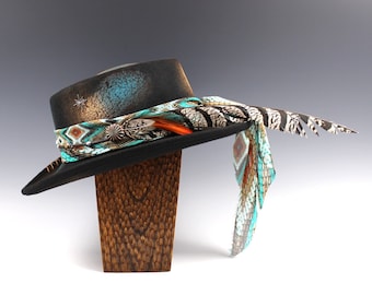 Puede incluir: Un sombrero negro con una banda decorativa con detalles turquesa, naranja y plateado. El sombrero tiene un adorno en forma de estrella y está adornado con una bufanda estampada y plumas. Descansa sobre un soporte de madera.