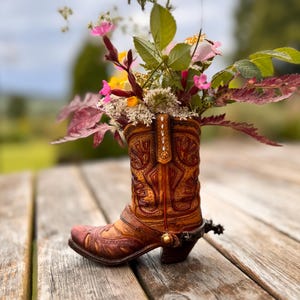 Peut inclure: Un vase en forme de botte de cowboy en cuir marron, rempli d'un arrangement floral coloré. La botte présente des motifs en relief détaillés et un éperon en métal. Les fleurs comprennent des fleurs roses, jaunes et blanches avec des feuilles vertes.