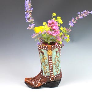 Peut inclure: Un vase décoratif en forme de botte de cowboy en céramique, marron avec des accents turquoise et dorés, rempli de fleurs colorées. La botte présente des détails complexes et est un objet de décoration unique.