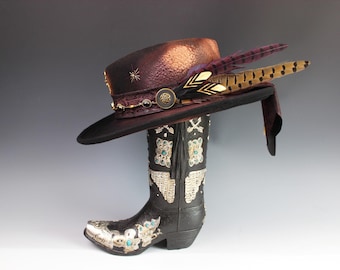 Puede incluir: Un sombrero decorativo de ala ancha, adornado con plumas y adornos, descansa sobre una bota de vaquero negra y plateada. El sombrero presenta una copa burdeos oscuro y una banda decorativa con detalles dorados. La bota está intrincadamente detallada con detalles plateados y turquesas.