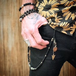 Può includere: Un set di braccialetti e collane di perline nere con dettagli di teschi e metallo. Una catena di cuoio con una moneta e un ciondolo a forma di punta di freccia pende dalla tasca. La persona indossa una camicia floreale e pantaloni neri.