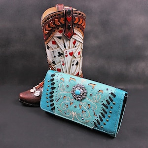 May include: A turquoise leather wallet with a decorative floral design and a large turquoise stone sits in front of a cowboy boot. The boot is brown and tan with playing card designs.