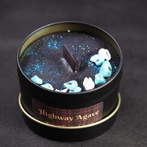 May include: A black candle in a round tin with a gold rim. The candle's surface is dark blue with blue glitter and small turquoise and white stones. The label reads "Highway Agave".