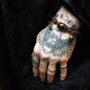 May include: Close-up of a hand adorned with rings and a wooden beaded bracelet. The hand features a large tattoo of a scarab beetle with outstretched wings. The background is a dark, textured fabric.