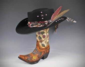 Puede incluir: Un sombrero de vaquero negro con adornos, incluyendo gemas rosas y una banda estampada, descansa sobre una bota de vaquero marrón y crema. La bota presenta diseños florales y una punta puntiaguda, creando una exhibición de temática occidental.