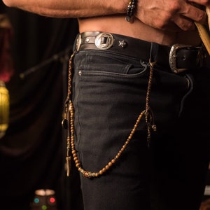 May include: A black leather belt with silver buckle and decorative studs. A beaded chain hangs from the belt loop. The person is holding drumsticks. The belt is worn with black jeans.