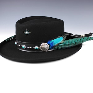 May include: Black fedora hat with a decorative band featuring turquoise stones and a silver emblem. The hat is adorned with a turquoise and green feather and a star design. The hat has a 5 cm brim.
