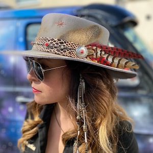 May include: A grey fedora hat with a snakeskin band, red and brown feathers, and a silver concho. The hat has a distressed look with a gold-toned brim and is adorned with red star-shaped embellishments. The hat is worn by a person wearing sunglasses.