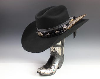 Puede incluir: Un sombrero de vaquero negro con una banda decorativa y detalles de plumas, colocado sobre una bota de vaquero negra y plateada. El sombrero presenta una banda decorativa con adornos plateados. La bota tiene intrincados diseños plateados y turquesas.
