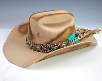 Puede incluir: Un sombrero de vaquero color beige con una banda decorativa de piel de serpiente, detalles turquesa y plumas coloridas. El sombrero presenta un diseño de estrella en la corona y un ala ancha. Un elegante accesorio western.