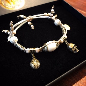 May include: A handcrafted bracelet featuring a cream-colored central bead and smaller wooden beads. The bracelet includes metal accents, a skull charm, and a decorative pendant. The bracelet is displayed on a black surface.