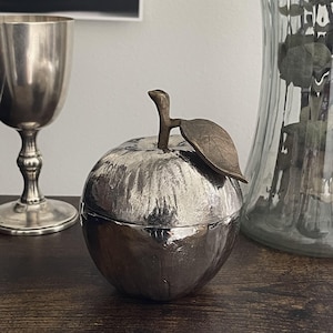 May include: A silver-toned apple-shaped decorative object with a textured, metallic finish. The piece features a distinct stem and a single leaf attached to the top, both rendered in a darker, bronze-toned metal, creating a contrast in colour.