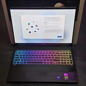 May include: A black laptop with a rainbow backlit keyboard is open, showing a Microsoft account setup screen. The laptop has a large screen and a touchpad. Intel and NVIDIA stickers are visible on the bottom right.