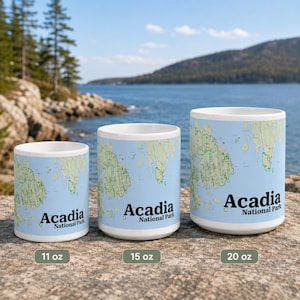 May include: Three white ceramic mugs of varying sizes, labeled 11 oz, 15 oz, and 20 oz. Each mug features a light blue and green topographical map print of Acadia National Park with the black text Acadia National Park printed on the front.