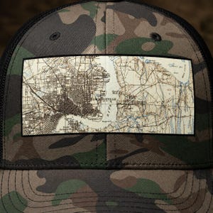 May include: A camouflage print trucker hat featuring a rectangular patch on the front. The patch displays a vintage-style topographic map of Jacksonville, Florida, with detailed street grids, river paths, and the text Jacksonville printed clearly.