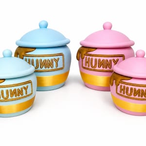 May include: Four ceramic jars with lids, two in light blue and two in light pink. Each jar features a gold band at the base and a raised brown rectangular label with the word HUNNY in relief, accompanied by a sculpted brown honey drip effect.