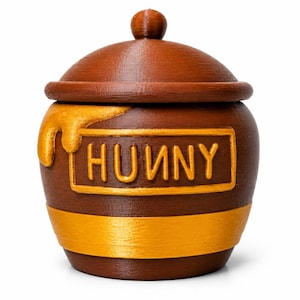 Op de afbeelding: Een bruine honingpot met een afneembaar deksel en een knop aan de bovenkant. De pot heeft een rechthoekig label met het woord HUNNY in verhoogde gele letters. Een geel druipeffect is zichtbaar aan de linkerkant en een gele band omcirkelt de basis.