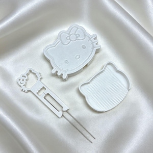 May include: A white plastic Hello Kitty cookie cutter set consisting of a character-shaped outline cutter, a detailed face stamper, and a long, thin metal-pronged tool for removing dough from the intricate sections of the cutter design.