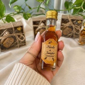 May include: A small glass bottle filled with amber liquid, featuring a gold cap and a label with the names "Nicola & Jennifer" and the date 02.05.2026. Several similar bottles and gift boxes are in the background, suggesting a wedding favor or gift.