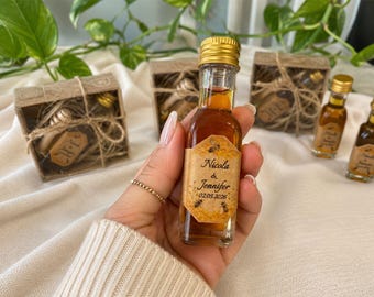 May include: A miniature glass bottle filled with amber liquid, with a gold cap and a label bearing the names "Nicola & Jennifer" and the date 02.05.2026. The bottle is held in a hand, with other bottles and gift boxes in the background.