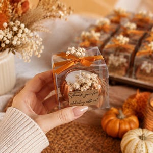 Può includere: Una scatola regalo quadrata trasparente contenente una candela color rame a forma di zucca, decorata con fiori bianchi secchi e un nastro color arancio bruciato. Una piccola etichetta rettangolare riporta il testo Thank you for celebrating with us.