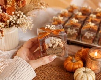 May include: A clear square gift box containing a copper-coloured pumpkin-shaped candle decorated with dried white flowers and a burnt orange ribbon. A small rectangular tag attached to the front features the text Thank you for celebrating with us.