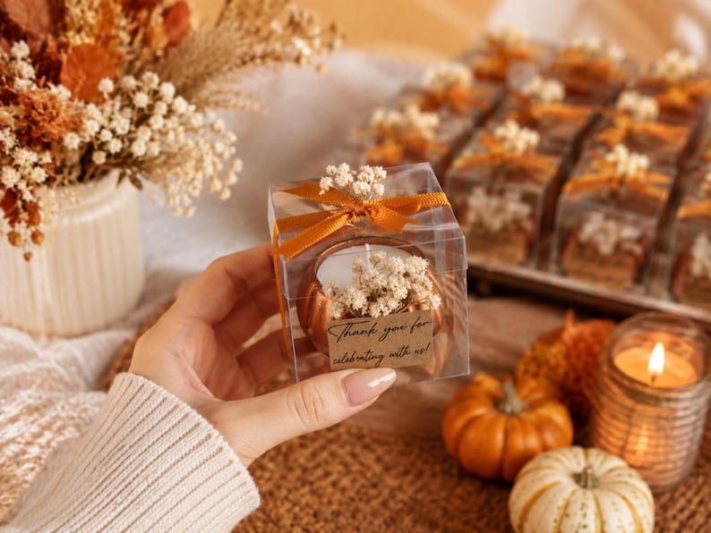 May include: A clear square gift box containing a copper-colored pumpkin-shaped candle decorated with dried white flowers and a burnt orange ribbon. A small rectangular tag attached to the front features the text Thank you for celebrating with us.