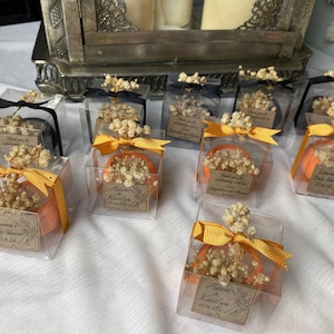 May include: Clear square plastic favor boxes containing small orange pumpkin-shaped items topped with dried baby's breath flowers. Each box is tied with a gold or black ribbon and features a rectangular paper tag with printed text and dates.