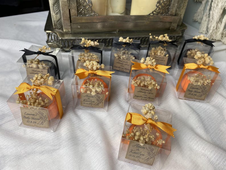 May include: Clear square plastic favor boxes containing small orange pumpkin-shaped items topped with dried baby's breath flowers. Each box is tied with a gold or black ribbon and features a rectangular paper tag with printed text and dates.