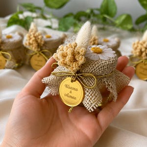 May include: Small glass jars wrapped in burlap, adorned with dried flowers, a daisy, and a gold tag. The jars are tied with gold string. The tag reads "Usama & Asmae".