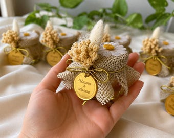 May include: Small glass jars wrapped in hessian, decorated with dried flowers, a daisy, and a gold tag. The jars are tied with gold string. The tag reads "Usama & Asmae".