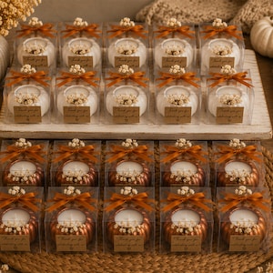 May include: A collection of pumpkin-shaped candles in clear square boxes, arranged in two rows. The top row features white pumpkin candles, while the bottom row contains metallic copper pumpkin candles. Each box is tied with a brown ribbon and a tag.