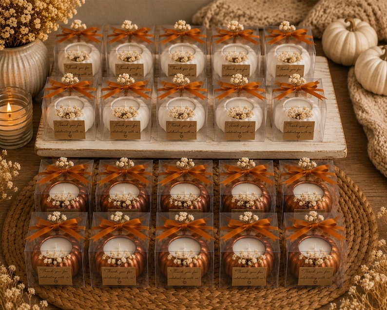 May include: A collection of pumpkin-shaped candles in clear square boxes, arranged in two rows. The top row features white pumpkin candles, while the bottom row contains metallic copper pumpkin candles. Each box is tied with a brown ribbon and a tag.