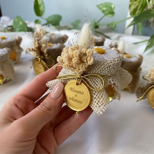 Op de afbeelding: Kleine glazen potjes gevuld met een gouden vloeistof, waarschijnlijk honing, zijn versierd met jute, gedroogde bloemen en madeliefjes. Elk potje heeft een gouden label met de namen "Usama & Asmae" gegraveerd. De potjes zijn op een wit oppervlak geplaatst.