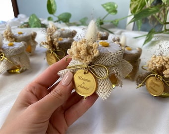 May include: Small glass jars filled with a golden liquid, possibly honey, are decorated with burlap, dried flowers, and daisy accents. Each jar has a gold tag with the names "Usama & Asmae" engraved on it. The jars are arranged on a white surface.