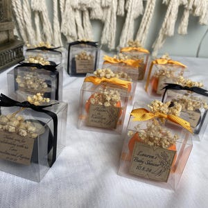 May include: A collection of clear square gift boxes containing small items, each topped with dried white flowers and a ribbon. Some boxes feature black ribbons, others orange. Each box includes a small brown tag with printed text and dates.