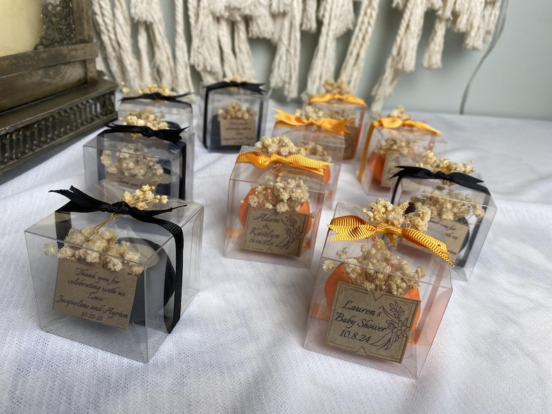 May include: A collection of clear square gift boxes containing small items, each topped with dried white flowers and a ribbon. Some boxes feature black ribbons, others orange. Each box includes a small brown tag with printed text and dates.