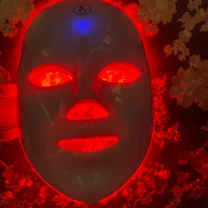 May include: A white LED light therapy mask with red lights illuminating the eye, nose, and mouth areas. The mask has a blue light on top. The background is a dark color with a floral pattern.