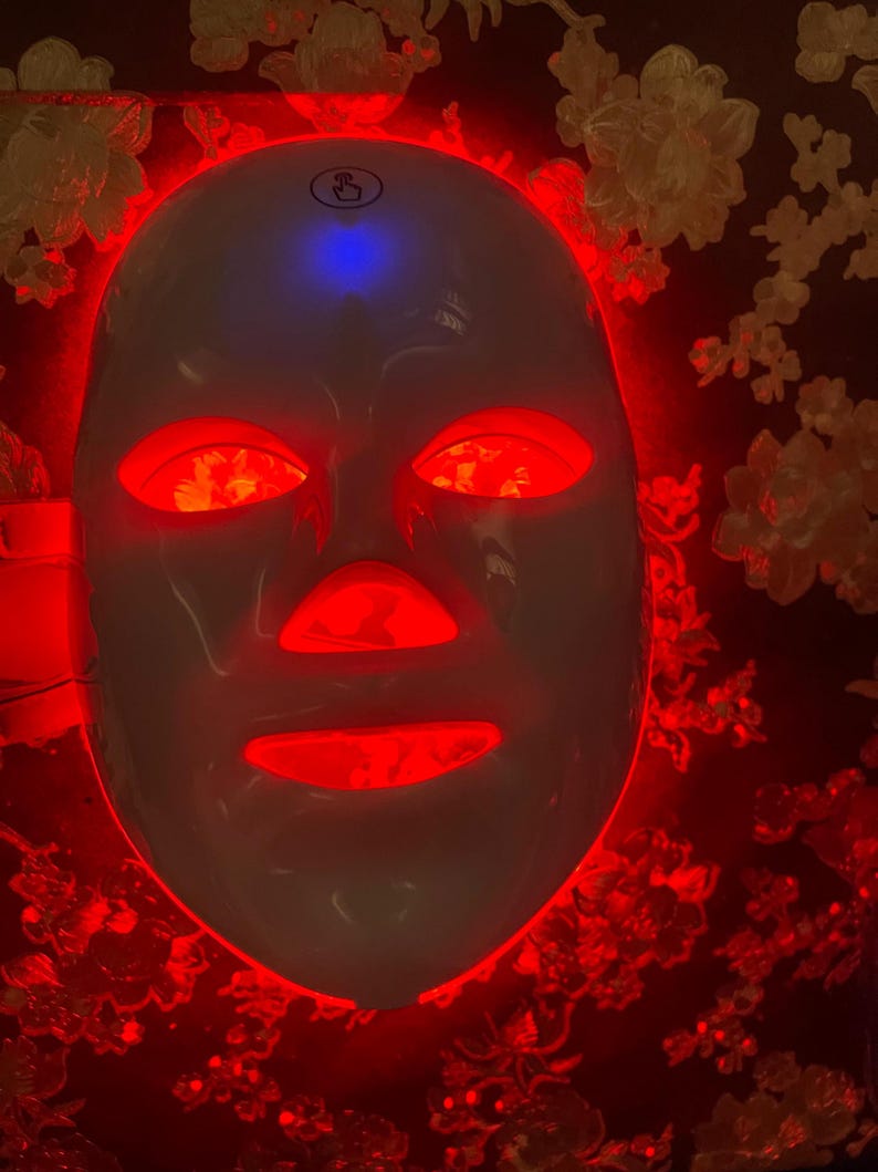 May include: A white LED light therapy mask with red lights illuminating the eye, nose, and mouth areas. The mask has a blue light on top. The background is a dark color with a floral pattern.