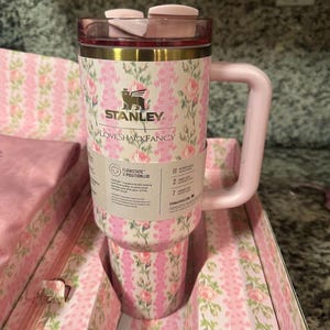 Może przedstawiać: R&oacute;żowo-biały kubek Stanley LoveShackFancy w kwiatowy wz&oacute;r z pasującym r&oacute;żowym uchwytem i pokrywką. Kubek posiada etykietę FlowState 3-position lid oraz tekst marki. Jest prezentowany w pasującym kwiatowym pudełku z r&oacute;żowym materiałem.