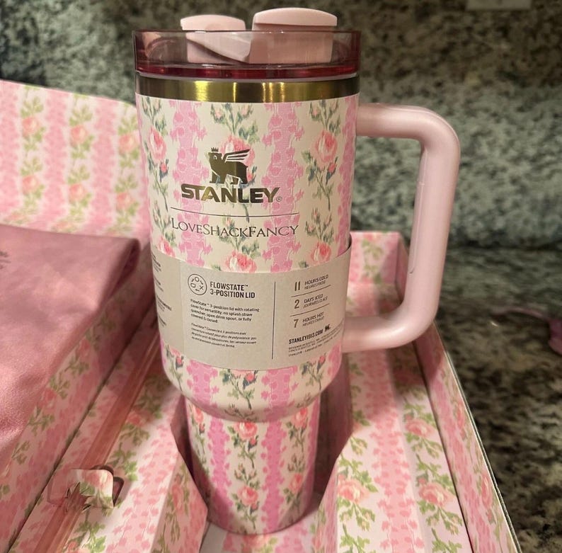 Może przedstawiać: R&oacute;żowo-biały kubek Stanley LoveShackFancy w kwiatowy wz&oacute;r z pasującym r&oacute;żowym uchwytem i pokrywką. Kubek posiada etykietę FlowState 3-position lid oraz tekst marki. Jest prezentowany w pasującym kwiatowym pudełku z r&oacute;żowym materiałem.