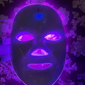 May include: A white LED light therapy mask with a glowing purple interior and exterior light. The mask has openings for the eyes, nose, and mouth, and is designed to fit the face. The background is a dark floral pattern.