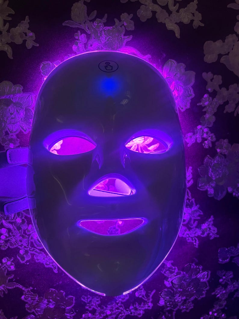 May include: A white LED light therapy mask with a glowing purple interior and exterior light. The mask has openings for the eyes, nose, and mouth, and is designed to fit the face. The background is a dark floral pattern.