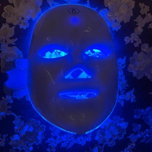 May include: A white LED light therapy mask with blue illumination around the eyes, nose, and mouth. The mask is designed to cover the entire face. The background is a dark blue floral pattern.